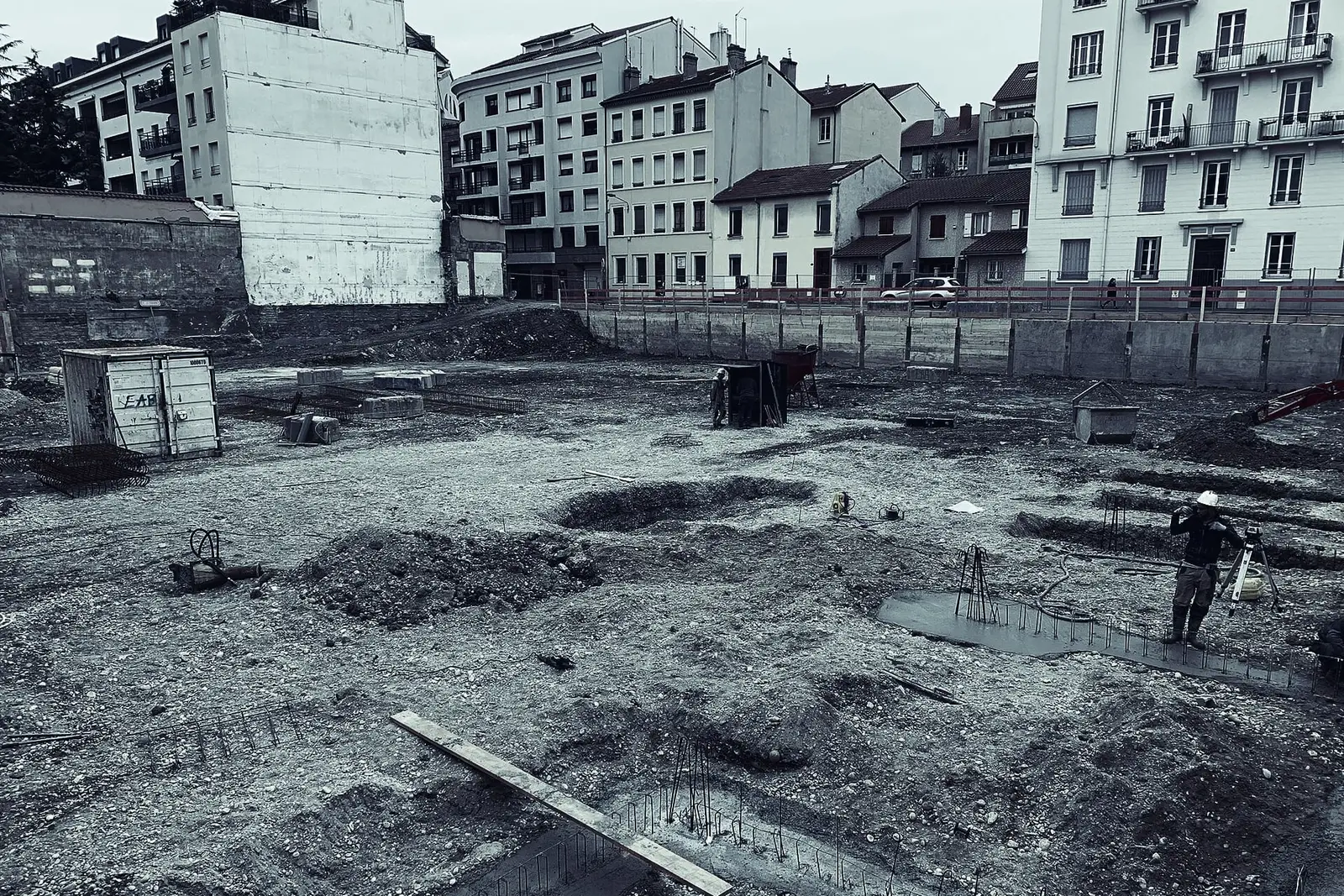 Terrain de chantier en ville au stade des fondations : sol terrassé, semelles coulées et fers d'attente, un ouvrier au fond, des immeubles tout autour.