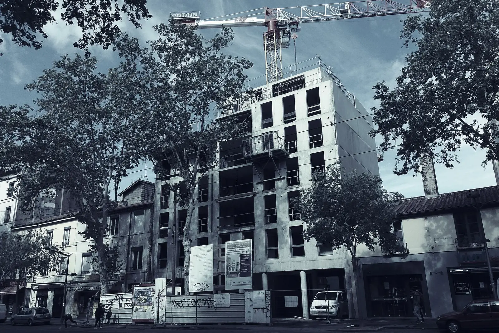 Immeuble de logements en construction dans une rue commerçante, structure en béton sur plusieurs étages, grue à tour au-dessus et palissade au pied.