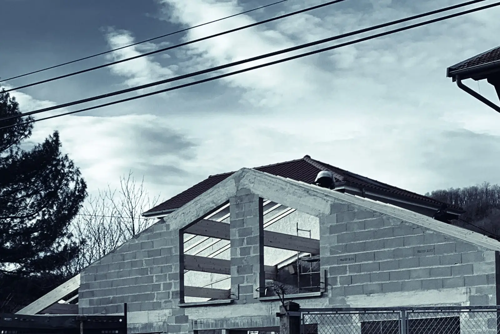 Maison individuelle en construction : murs en parpaings montés, fermes de charpente en bois posées, à côté d'une maison habitée aux volets en bois.