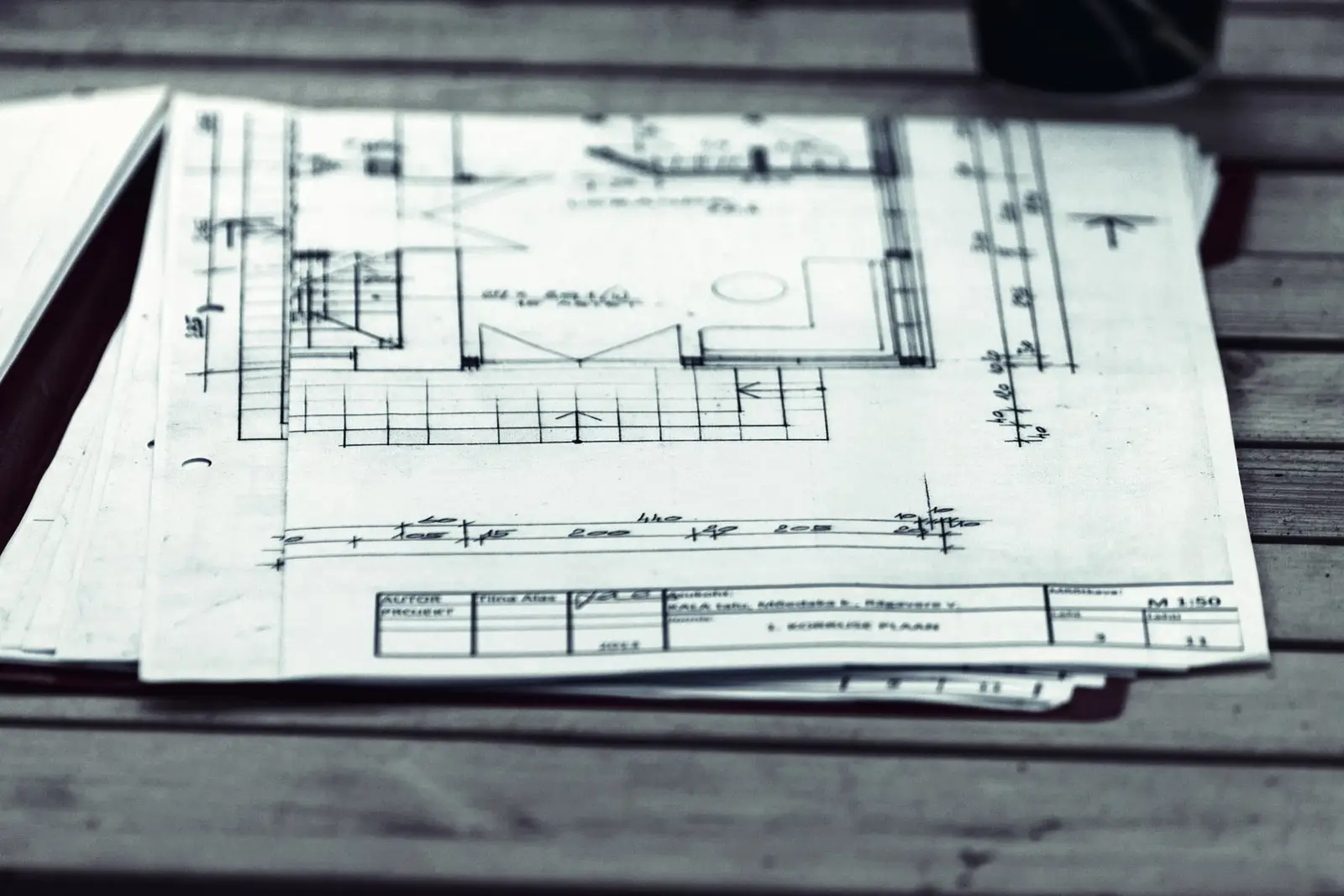 Liasse de plans imprimés posée sur une table en bois, avec le cartouche et les cotes visibles en bas de la première feuille.