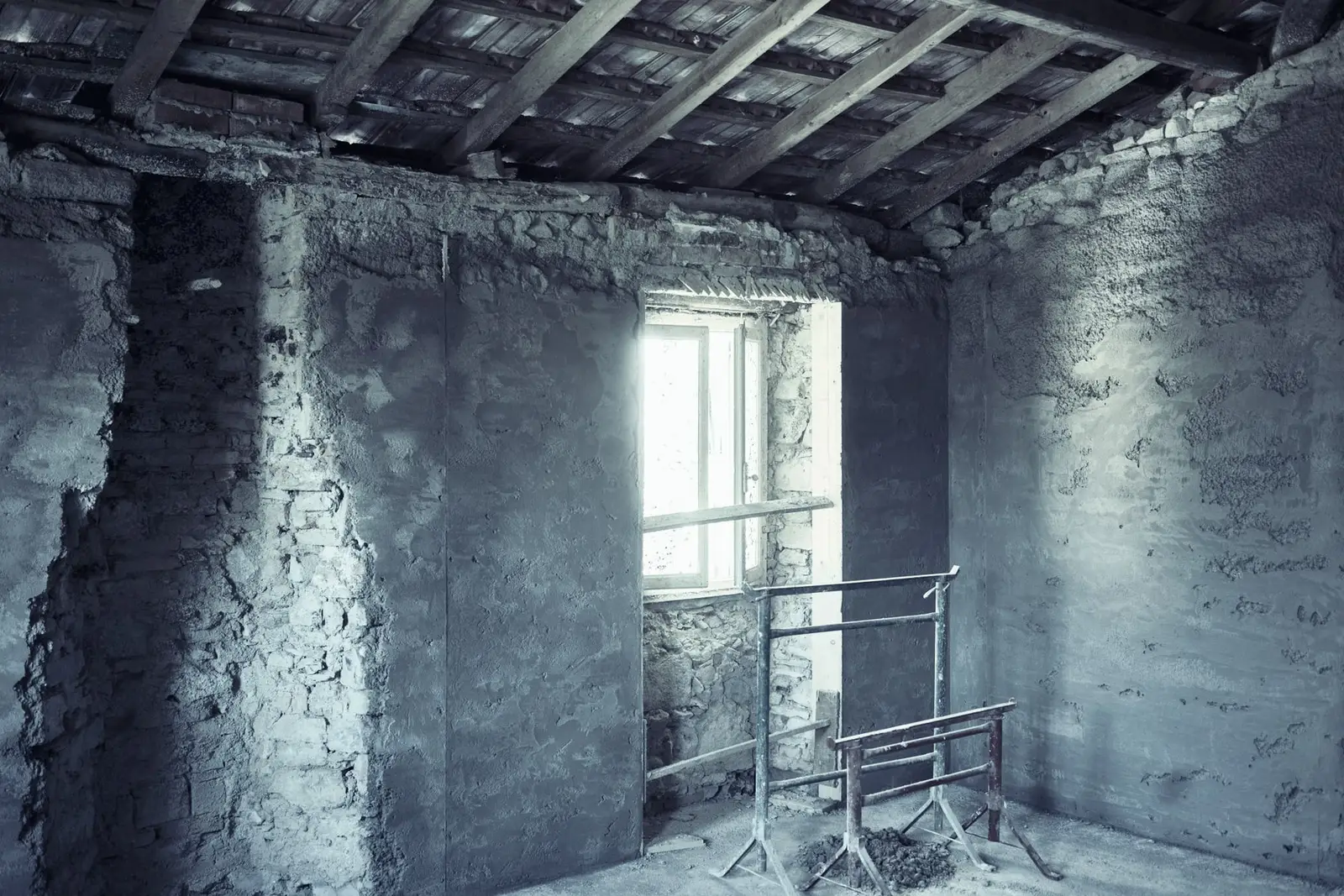 Pièce d'une maison ancienne en pleine rénovation : murs de pierre en partie enduits, charpente apparente, tréteaux devant la fenêtre.